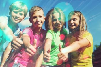 Czworo dzieci stoi na zewnątrz, uśmiechając się i trzymając kciuki. Ubrane w kolorowe koszulki, cieszą się światłem słonecznym z efektem flary obiektywu, uchwycając radosny nastrój w placówce wsparcia dziennego na Mokotowie.