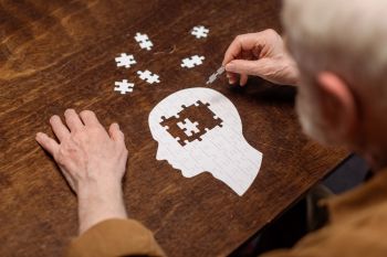 starszy mężczyzna siedzi przy stole i układa białe puzzle w kształcie głowy ludzkiej, metafora choroby alzheimera