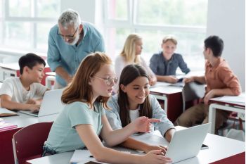 Dwie dziewczyny siedzące przy biurku w klasie pracują razem na laptopie, uśmiechając się. Inni uczniowie i nauczyciel są w tle, również zaangażowani w zajęcia grupowe. Klasa jest jasna i nowoczesna.