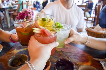 Cztery osoby brzęczą kolorowymi koktajlami przy restauracyjnym stole zastawionym miskami i talerzami, świętując w żywej, swobodnej atmosferze.