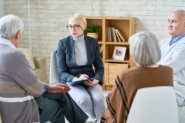 Kobieta o krótkich blond włosach i w okularach, trzymająca tablicę, prowadzi grupę wsparcia dla doświadczających uzależnień z trzema starszymi dorosłymi siedzącymi w kręgu w przytulnym, dobrze oświetlonym pokoju wyłożonym półkami.