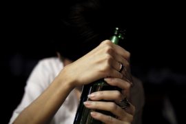 Zrozpaczona kobieta ze spuszczoną głową, mocno ściskająca zieloną szklaną butelkę obiema rękami na ciemnym tle. Jej twarz jest zasłonięta, odzwierciedlając zmagania alkohol a kobieta.