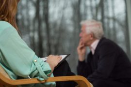 Kobieta trzyma schowek i słucha, podczas gdy starszy mężczyzna siedzi naprzeciwko niej, wyglądając na zamyślonego i wpatrując się w okno z drzewami na zewnątrz. Sceneria sugeruje sesję doradczą skoncentrowaną na kwestiach takich jak alkoholizm.