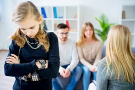 Nastoletnia dziewczyna stoi ze skrzyżowanymi ramionami, patrząc w dół i wyglądając na zdenerwowaną, podczas gdy troje dorosłych - prawdopodobnie dla rodzin dotkniętych problemem uzależnienia od alkoholu - siedzi na kanapie w tle, obserwując ją z niepokojem w jasnym salonie.