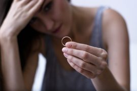 Kobieta o zatroskanym wyrazie twarzy trzyma pierścionek w dłoni, opierając drugą rękę na czole, co sugeruje uczucie smutku lub kontemplacji, prawdopodobnie związane ze współuzależnieniem od alkoholu.