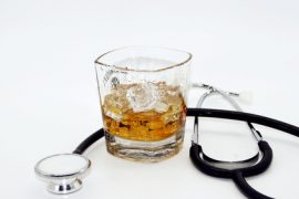 Szklanka whisky z lodem stoi obok czarnego stetoskopu na białym tle, subtelnie podkreślając szkody zdrowotne picia alkoholu.