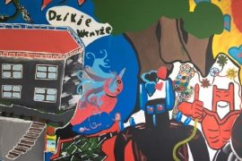 Kolorowy, chaotyczny mural stworzony jako projekt dla młodzieży przedstawia budynek szkoły, abstrakcyjne kształty, zamaskowaną postać z rurkami, niebieskie serca, czerwonego superbohatera, drzewo i słowo "Akumulator" napisane pogrubionymi czerwonymi literami na żółtym tle.