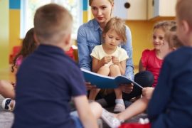 Kobieta zapewnia wsparcie dla dzieci, czytając książkę grupie małych dzieci siedzących w kręgu na podłodze w kolorowej klasie. Dzieci słuchają uważnie i wydają się zaangażowane.