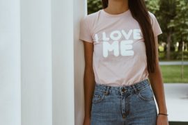 Osoba stoi na zewnątrz ubrana w jasnoróżową koszulkę z napisem "I LOVE ME" pogrubionymi białymi literami, w połączeniu z niebieskimi dżinsami z wysokim stanem, jakby była gotowa do wzięcia udziału w warsztatach miłości do siebie podczas konferencji DDA. Twarz nie jest widoczna na zdjęciu.