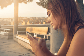 Młoda kobieta o brązowych włosach siedzi na zewnątrz na ławce, patrząc uważnie na swój smartfon - być może sprawdzając teleporady Karan - gdy zachodzi słońce, rzucając ciepłe światło na drzewa i rozmazany krajobraz miasta w tle.