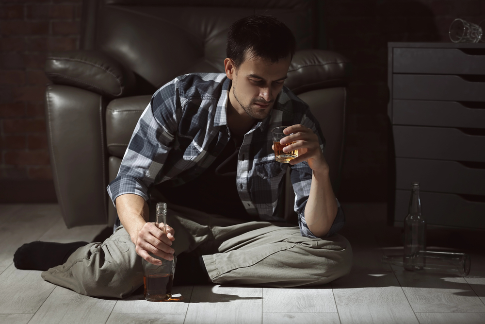 Mężczyzna siedzi na podłodze, trzymając w jednej ręce szklankę whisky, a w drugiej butelkę, i spogląda w dół z zamyśleniem. Tuż obok na podłodze leży pusta butelka.