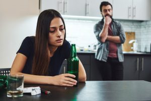 Młoda kobieta pije piwo przy stole, za nią stoi zatroskany mężczyzna