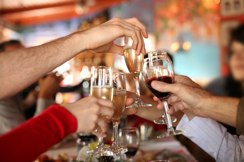 Kilka osób wznosi kieliszki z winem i szampanem, aby wznieść toast podczas uroczystego spotkania w pomieszczeniu; w tle widać rozmyte, kolorowe tło, a na stole widać potrawy.