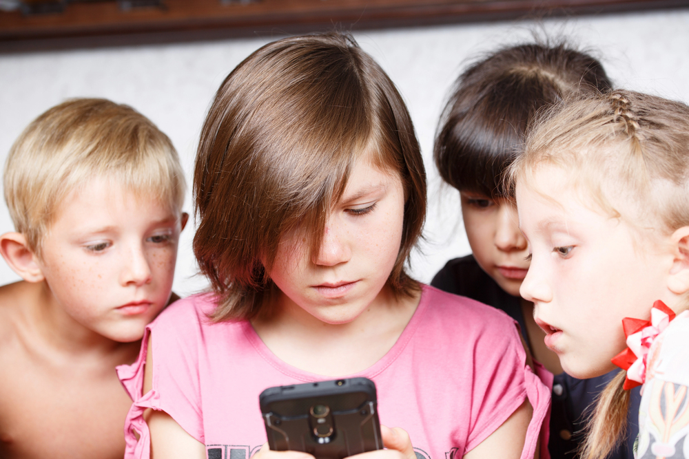 Czworo dzieci stoi blisko siebie, wpatrując się z uwagą w smartfon trzymany przez dziewczynkę w środku, ubraną w różową koszulkę. Wyglądają na skupione i zafascynowane tym, co dzieje się na ekranie.