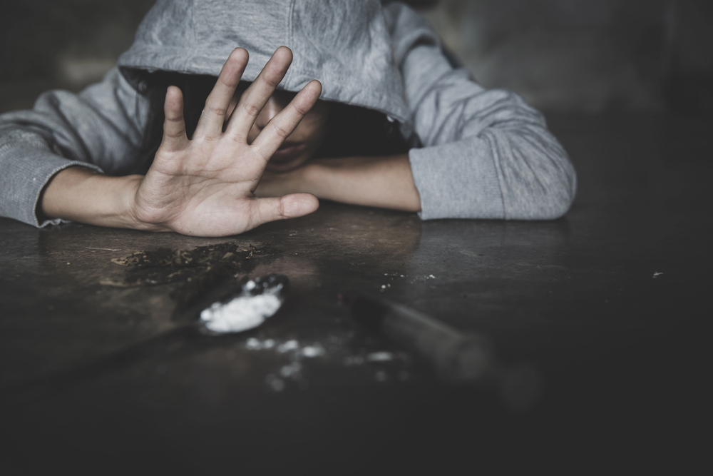 Osoba w szarej bluzie z kapturem siedzi przy stole z opuszczoną głową, zasłaniając twarz dłonią. Na stole widać akcesoria do zażywania narkotyków, w tym strzykawkę i proszek.