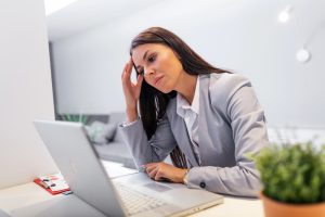 Kobieta w garniturze siedzi przy biurku, wyglądając na zestresowaną lub sfrustrowaną, wpatrując się w ekran laptopa. Opiera głowę na dłoni. Na biurku leży notes z klipsem oraz doniczkowa roślina.