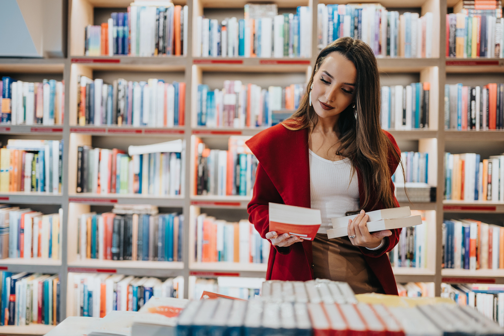 Kobieta w czerwonym płaszczu stoi w bibliotece lub księgarni, trzymając w rękach kilka książek i przeglądając okładkę jednej z nich, a w tle widać regały pełne książek.