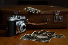 Zabytkowy aparat fotograficzny stoi na drewnianym stoliku przed zużytą brązową walizką, z kilkoma starymi czarno-białymi fotografiami rozrzuconymi na pierwszym planie, sugerując wspomnienia ukształtowane przez zmagania, takie jak uzależnienie od alkoholu.
