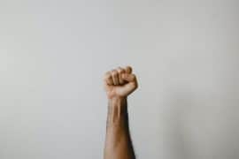 Uniesiona ręka z zaciśniętą pięścią na jednolitym jasnym tle, symbolizująca siłę, solidarność lub opór.