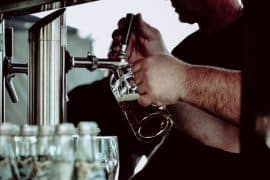 Osoba nalewa piwo z beczki z kranu do szklanego kufla, z innymi szklankami na pierwszym planie. Słabo oświetlona scena, skupiająca się na dłoniach barmana i kranie z piwem, subtelnie podkreśla codzienne rytuały związane z uzależnieniem od alkoholu.