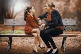 Kobieta i młoda dziewczyna siedzą na drewnianej ławce na zewnątrz, uśmiechając się i trzymając za ręce. Oboje noszą okulary i są ubrani w ciemne, jesienne ubrania. Tło jest rozmyte drzewami i miękkim, ciepłym oświetleniem.