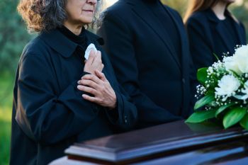Trzy osoby ubrane na czarno stoją na świeżym powietrzu przy trumnie udekorowanej białymi kwiatami. Jedna z nich przyciska chusteczkę do piersi, co sugeruje ponurą atmosferę pogrzebu.